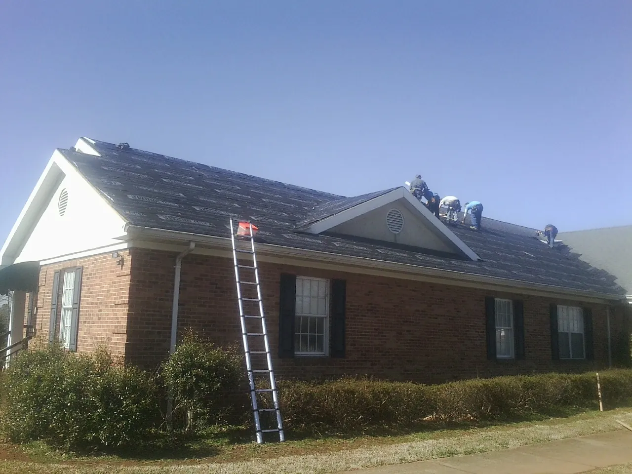 Crew tearing shingles off a brick ranch roof with a ladder set — Emergency Roof Repair work by our crew in Harvest
