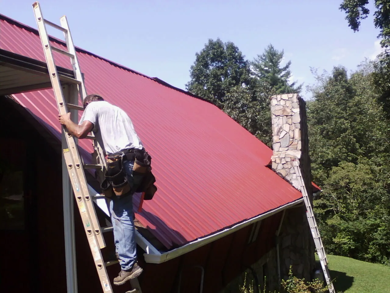 Roofer on a ladder beside a red metal roof and stone chimney — Soffit & Fascia Repair by Roofing Brothers in Harvest